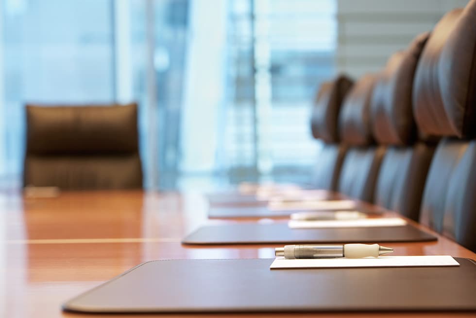 A conference room table set with leather writing pads, pens, and notepapers, with high-backed chairs arranged along the table and large windows in the background.