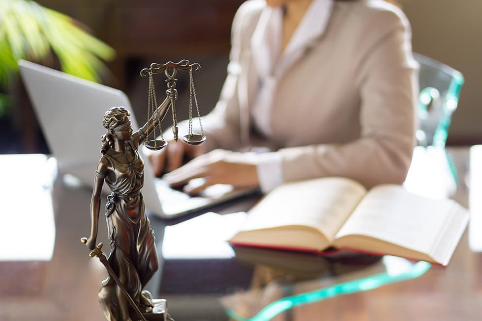 A statue of Lady Justice holding scales and a sword stands on a desk in the foreground, while a woman works on a laptop with an open book beside her in the background.