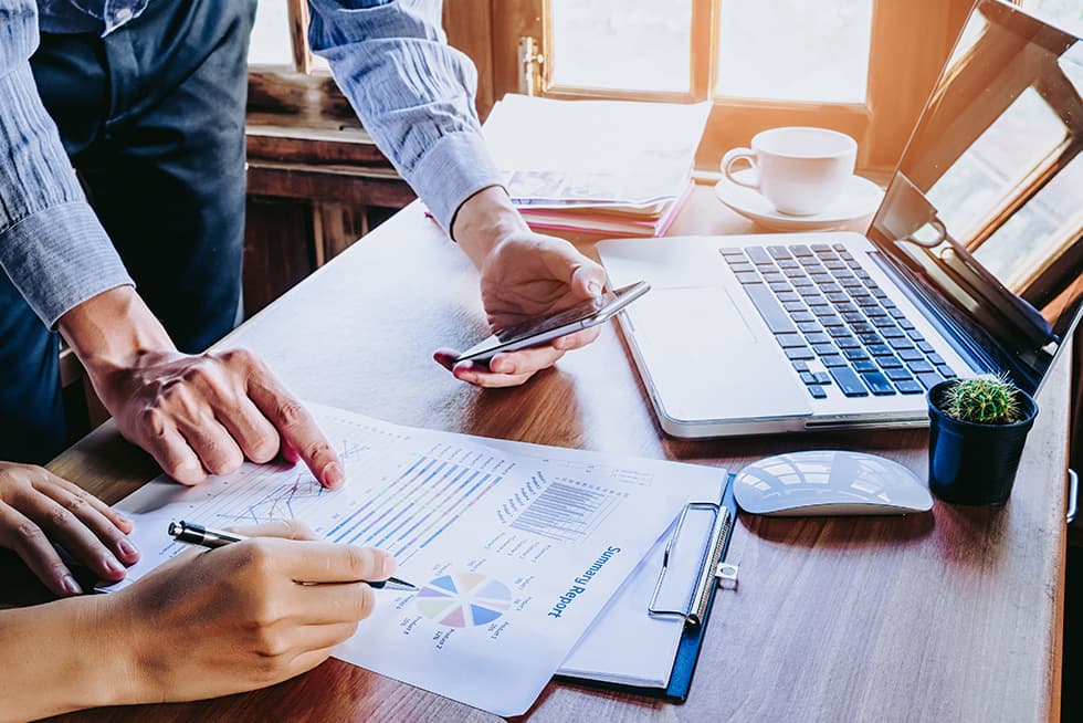 Two people reviewing a financial report at a desk, with one pointing at charts on a printed document while holding a smartphone, and a laptop, mouse, coffee cup, and small plant nearby.