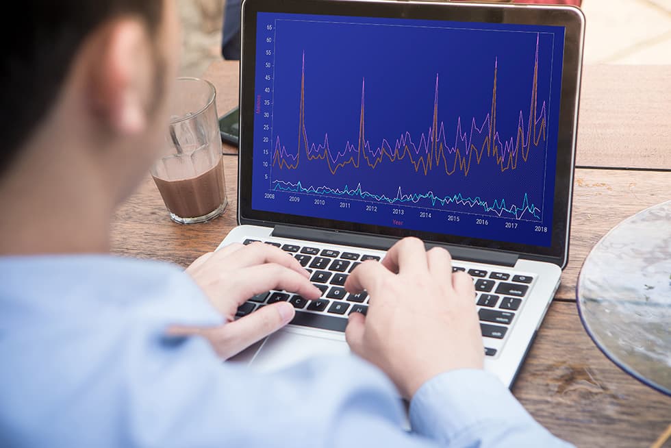 A person typing on a laptop displaying a multi-line financial chart on the screen, with a glass of chocolate drink placed beside the computer on a wooden table.