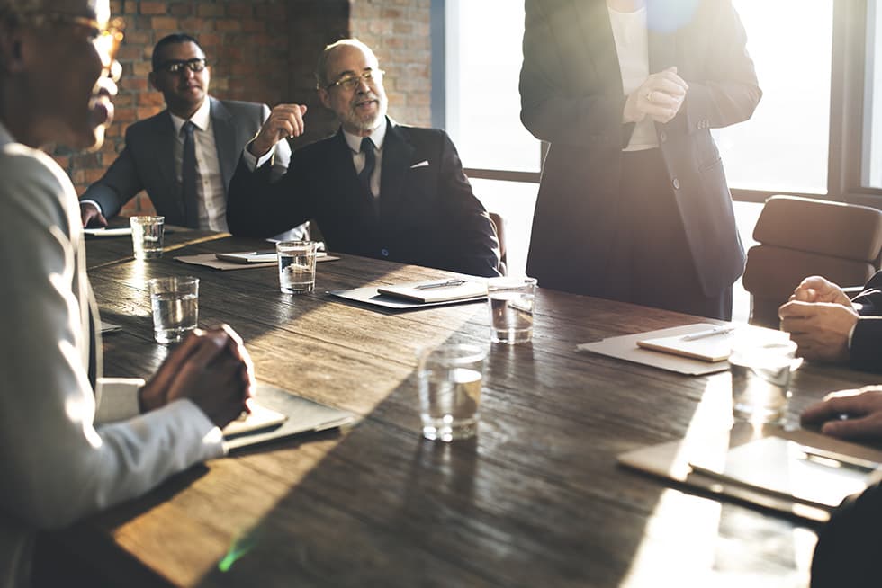 Several business professionals sit around a wooden conference table with notebooks and glasses of water, listening as one person stands and speaks during a meeting.