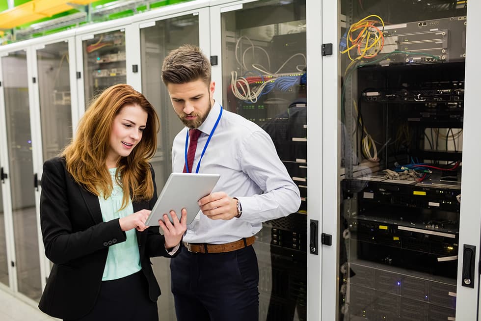 Two professionals stand in a server room in front of network racks, reviewing information on a tablet while cables and equipment are visible behind them.