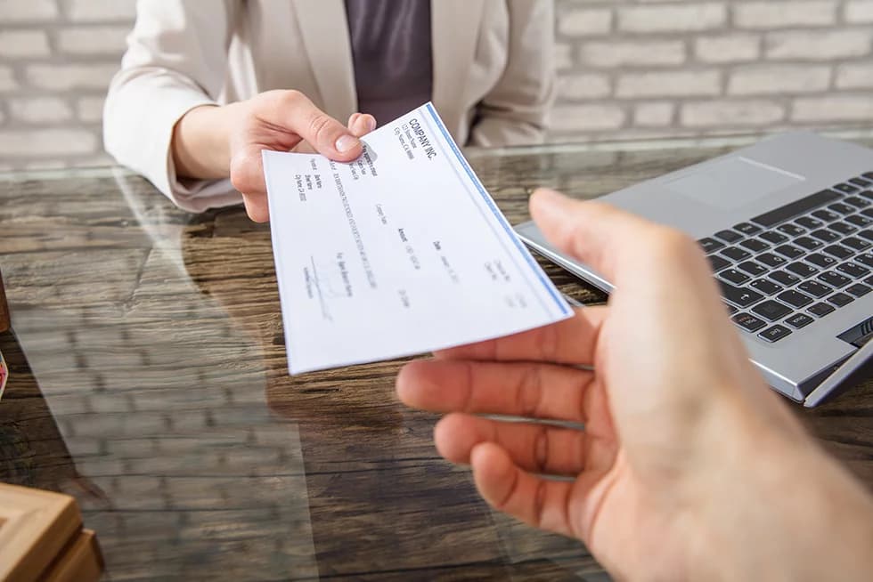 Close-up of a person handing over a check across a desk, with a laptop visible in the background.