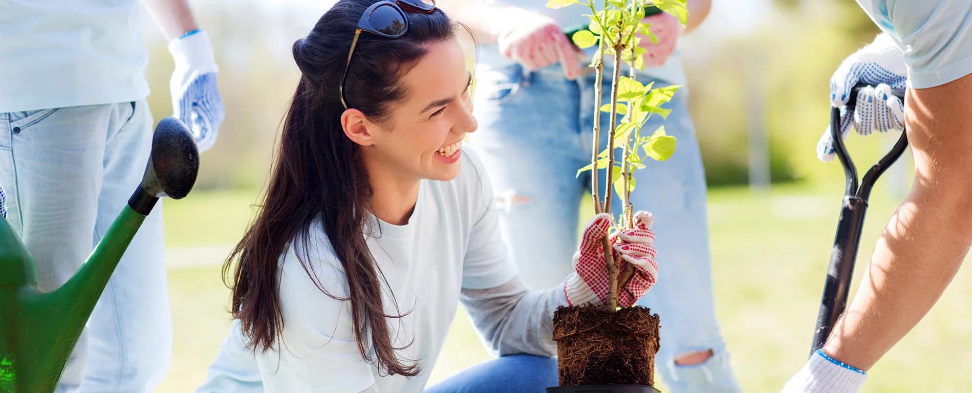 A smiling woman wearing gardening gloves holds a small tree sapling with soil around its roots while standing outdoors, as other people with gardening tools assist nearby.