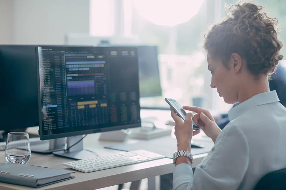 A woman sits at a desk in an office, looking at her smartphone while a large monitor beside her displays financial data and charts.
