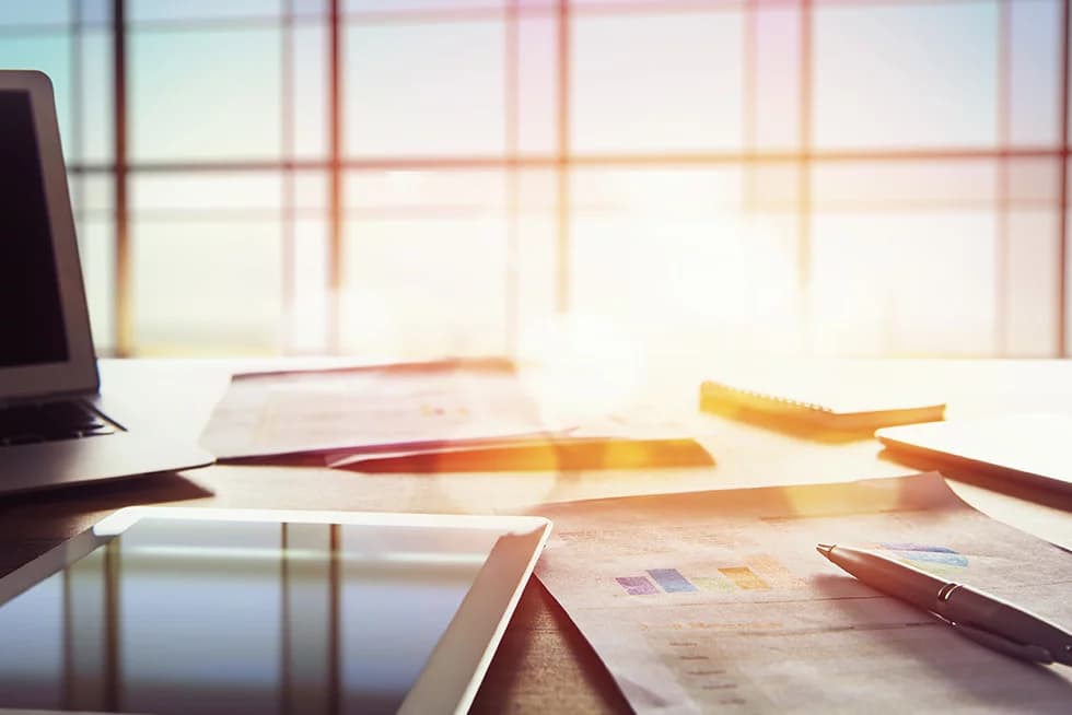 A sunlit desk with a laptop, tablet, printed charts, a notebook, and a pen, set in front of large windows with warm sunlight streaming in.