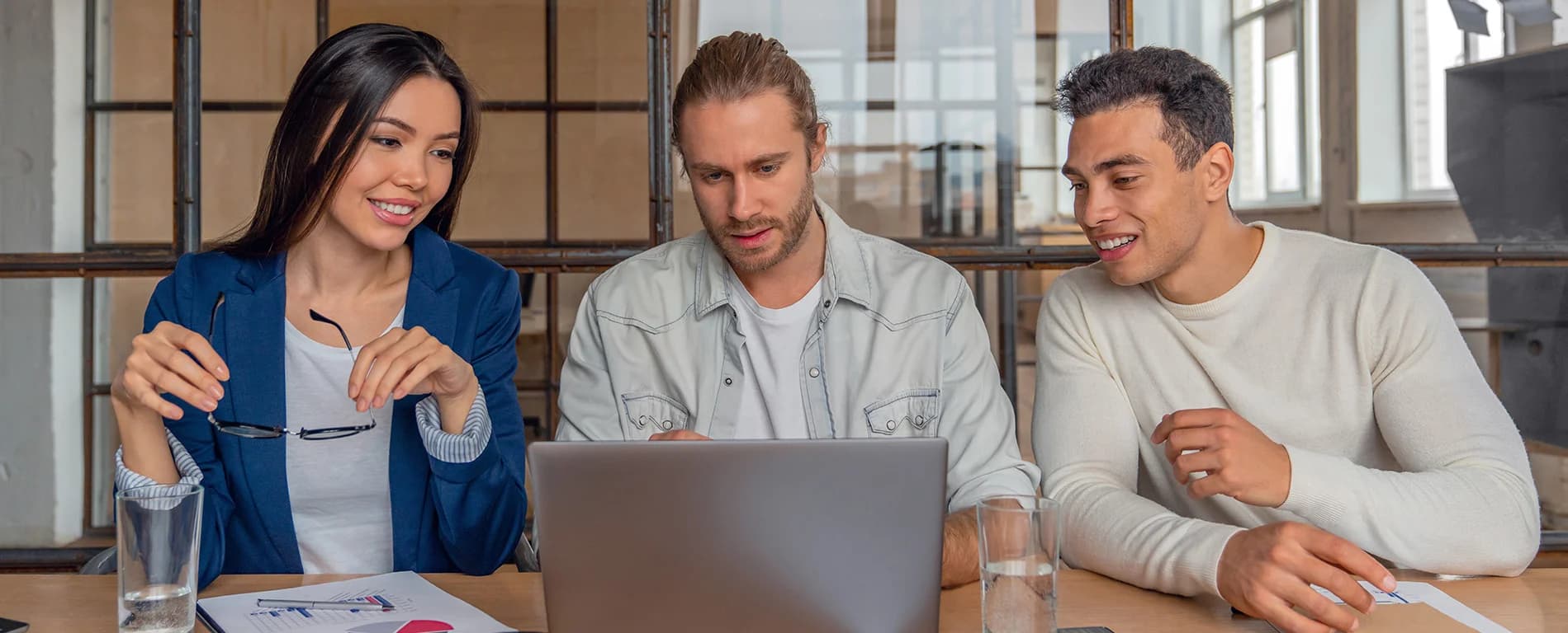 Three colleagues smiling and collaborating around a laptop, reviewing documents and data in a modern office setting.