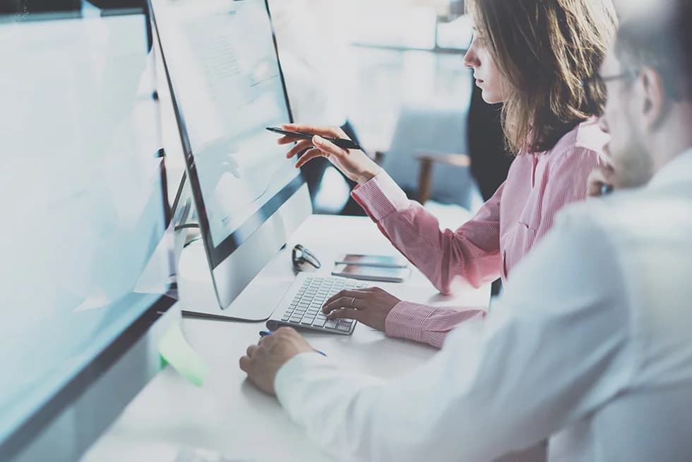 Two colleagues work together at a desk, with one pointing at data on a large computer screen using a pen while the other looks on attentively.