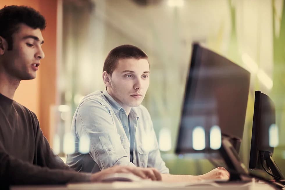 Two colleagues working together at a desktop computer, focused on the screen while discussing tasks in a modern office environment.