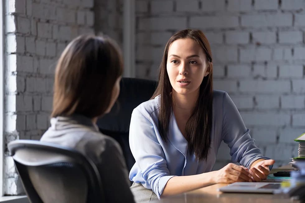 A woman in a light blue blouse speaks while sitting at a desk across from another woman in an office with a white brick wall background. A laptop and documents are on the table between them.