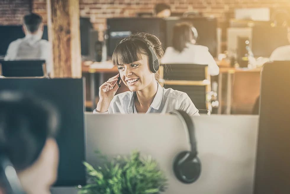 A woman wearing a headset sits at a desk in an office environment, smiling while talking on the phone. She is positioned behind computer monitors, with colleagues working at desks in the background. The setting appears to be a modern, open-plan workspace with warm lighting and exposed brick walls.