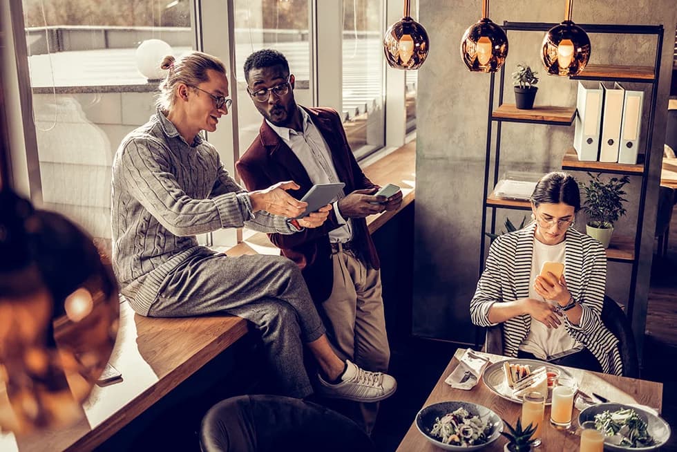 Three colleagues looking at a tablet in a modern café setting with food and drinks on the table.