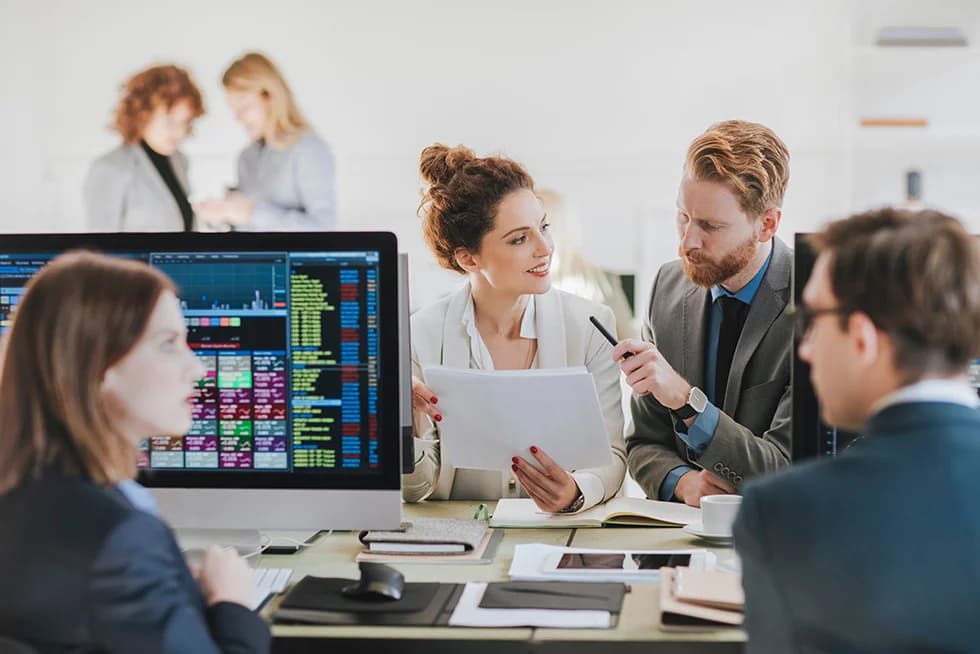 Team of professionals analyzing financial data on computer screens, discussing reports and reviewing documents in a modern office setting.