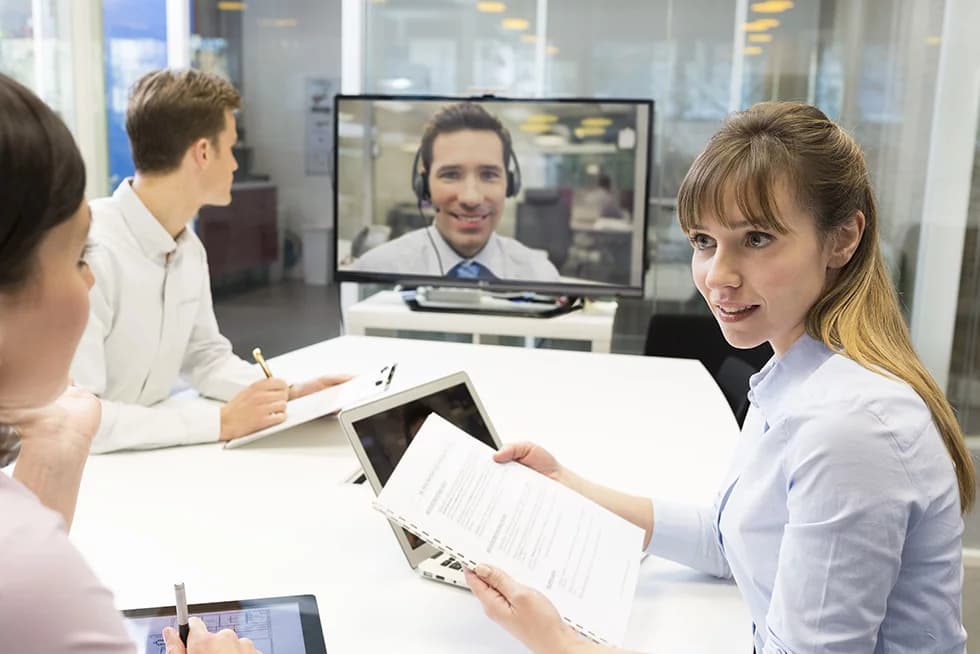 Three colleagues in a meeting room reviewing documents while a remote participant joins via video conference on a large screen.