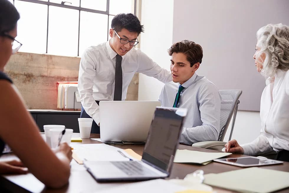 A group of colleagues sit around a table in a meeting, with one person standing and pointing at a laptop screen while others review documents and take notes.