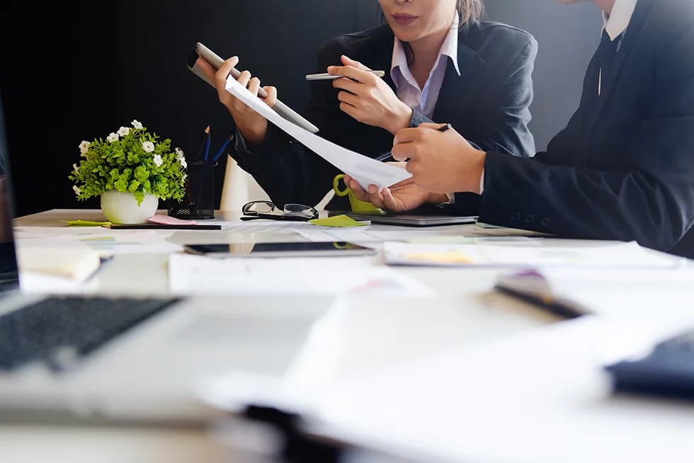 Two business professionals sit at a desk reviewing documents together, holding papers and a pen, with office supplies and a small potted plant nearby.