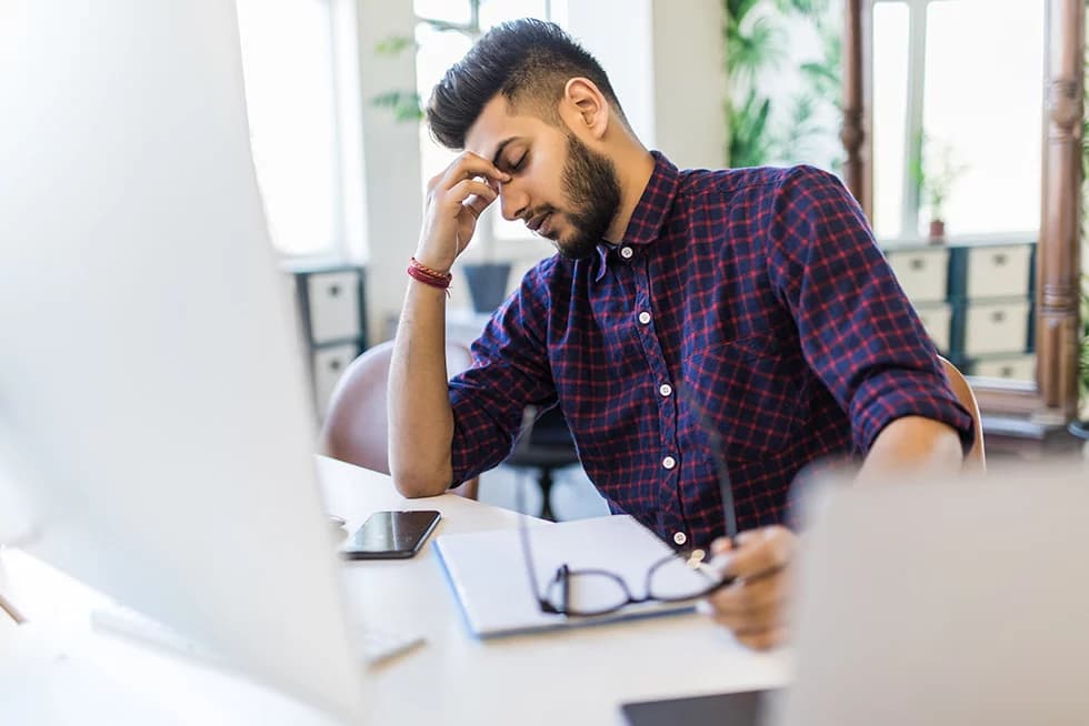 A young man sits at a desk looking stressed, holding his forehead while working on a computer, with a notebook, glasses, and smartphone nearby.