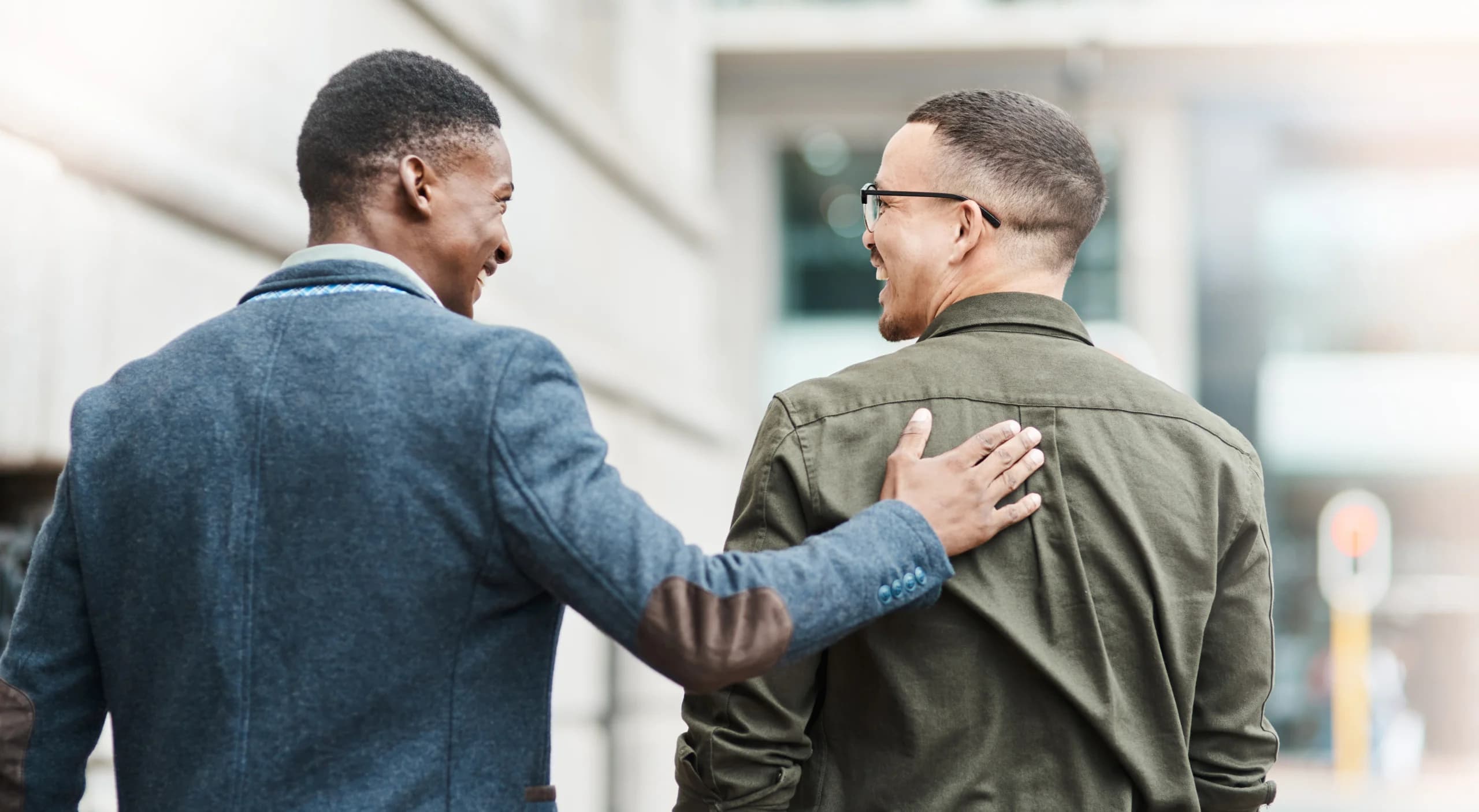 Two men walking together outdoors, smiling, with one patting the other on the back.