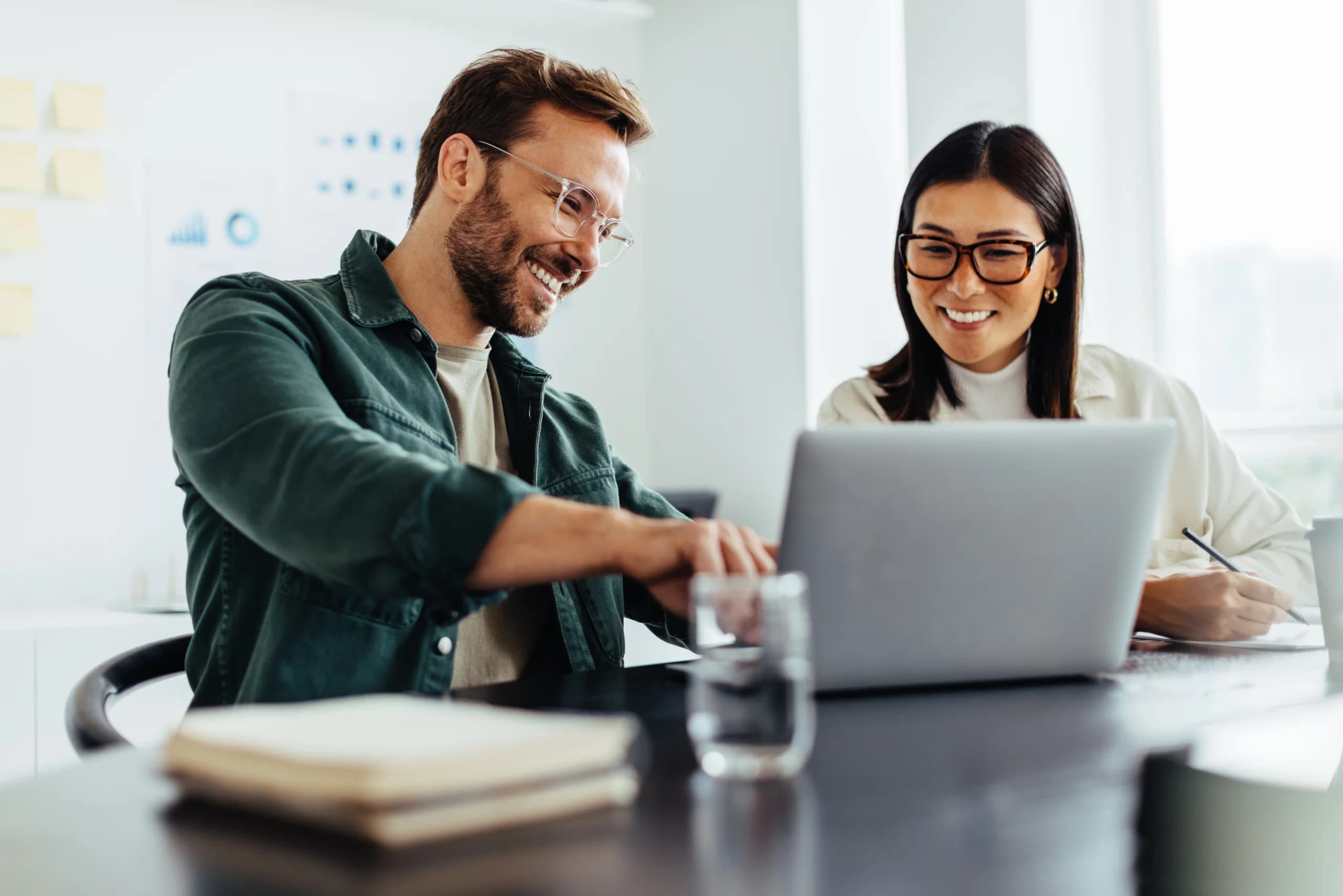 Two colleagues smiling while collaborating on a laptop during a meeting in a modern office.