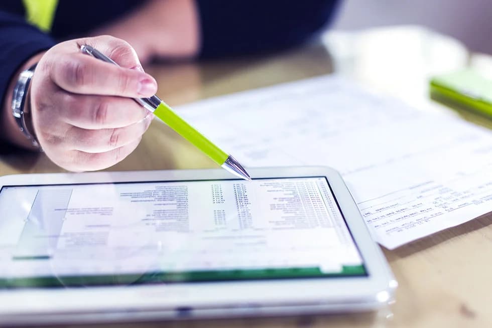 Close-up of a person holding a green pen and reviewing financial data on a tablet, with printed documents on a desk.