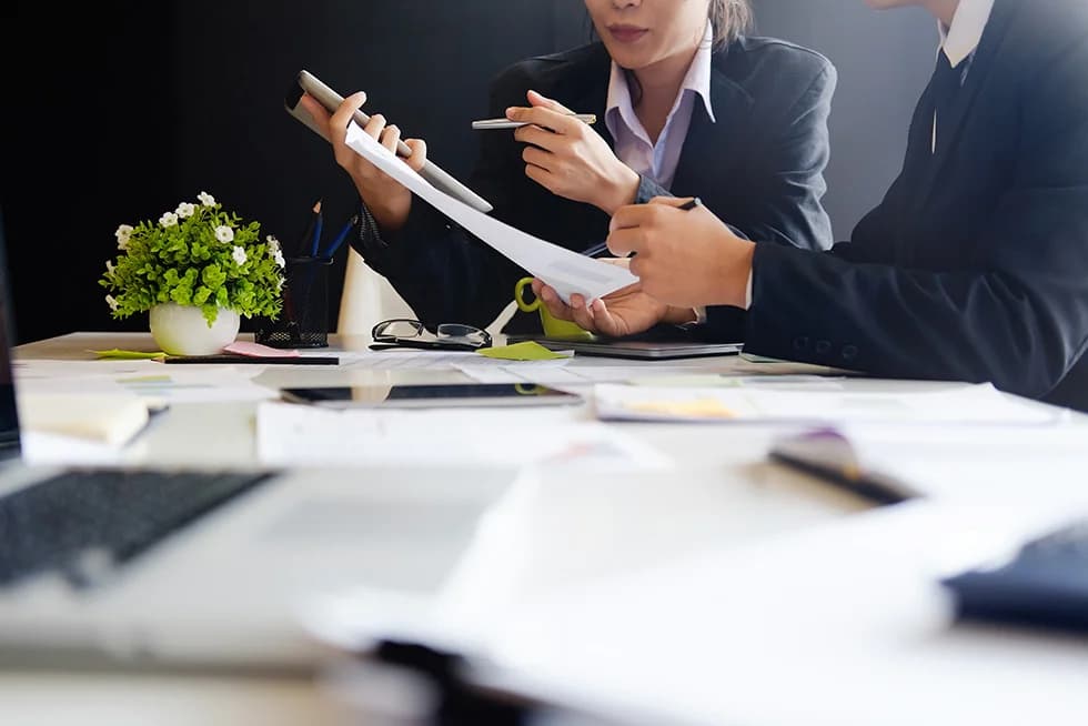 Two business professionals reviewing documents and discussing financial information at a desk with a calculator, laptop, and paperwork.