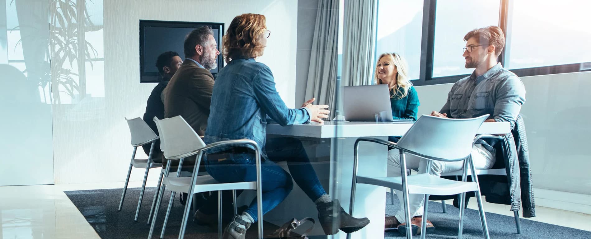 Four professionals sitting around a conference table in a modern office, discussing and collaborating during a team meeting with laptops and natural light from large windows.