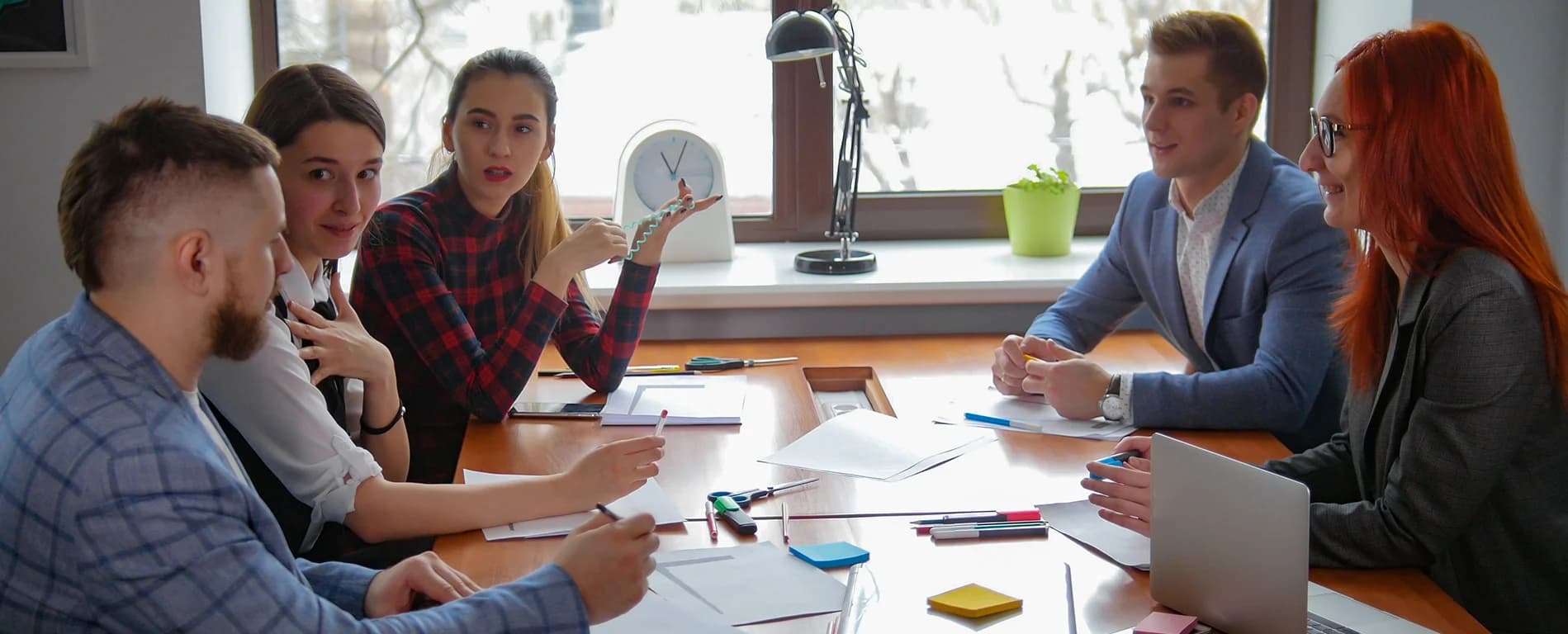Group of five colleagues gathered around a table in a bright office, discussing documents and collaborating during a team meeting.
