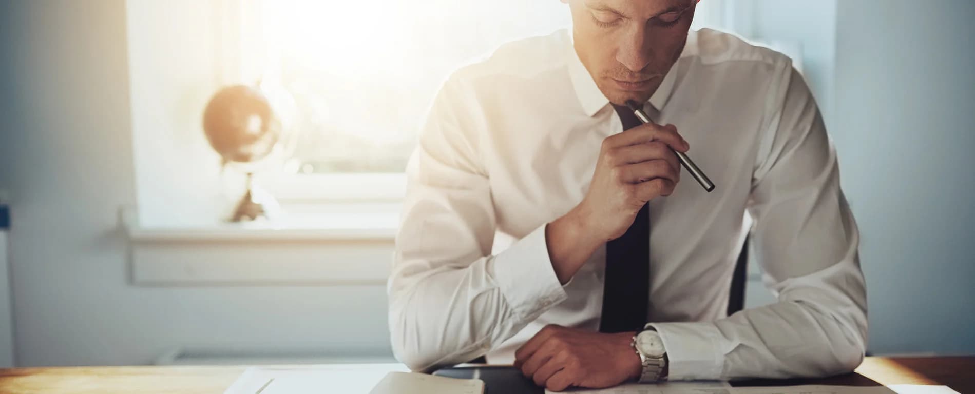 A businessman in a white shirt and tie sits at a desk, holding a pen to his lips while thoughtfully reviewing documents in warm sunlight.