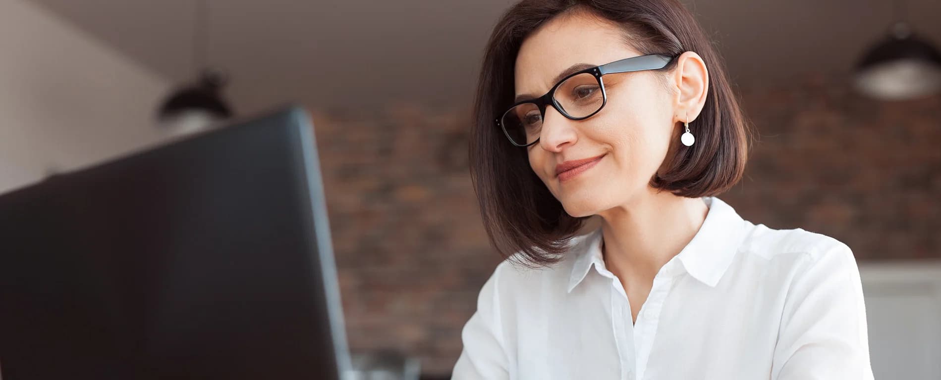 A woman wearing glasses and a white blouse smiles slightly while looking at a computer screen in a modern office setting.