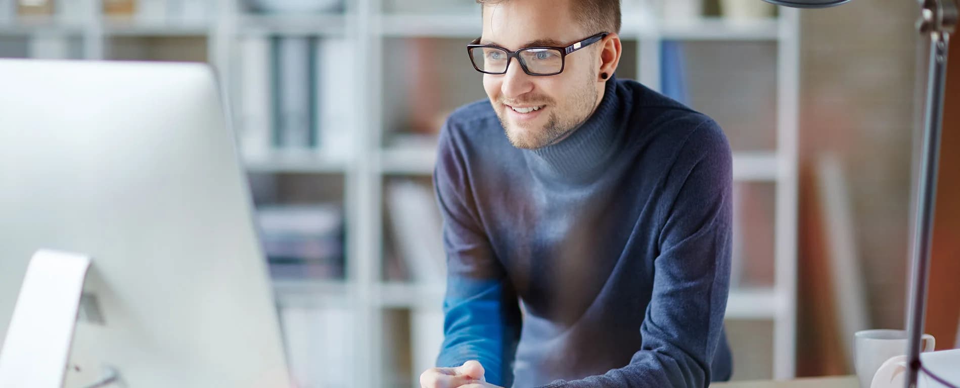 A man wearing glasses and a dark sweater leans forward and smiles while working at a desktop computer in a bright office setting.