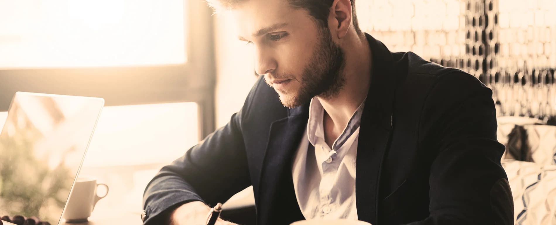 A man in a blazer sits at a table near a window, focused on writing or reviewing notes beside an open laptop and a cup of coffee in warm sunlight.