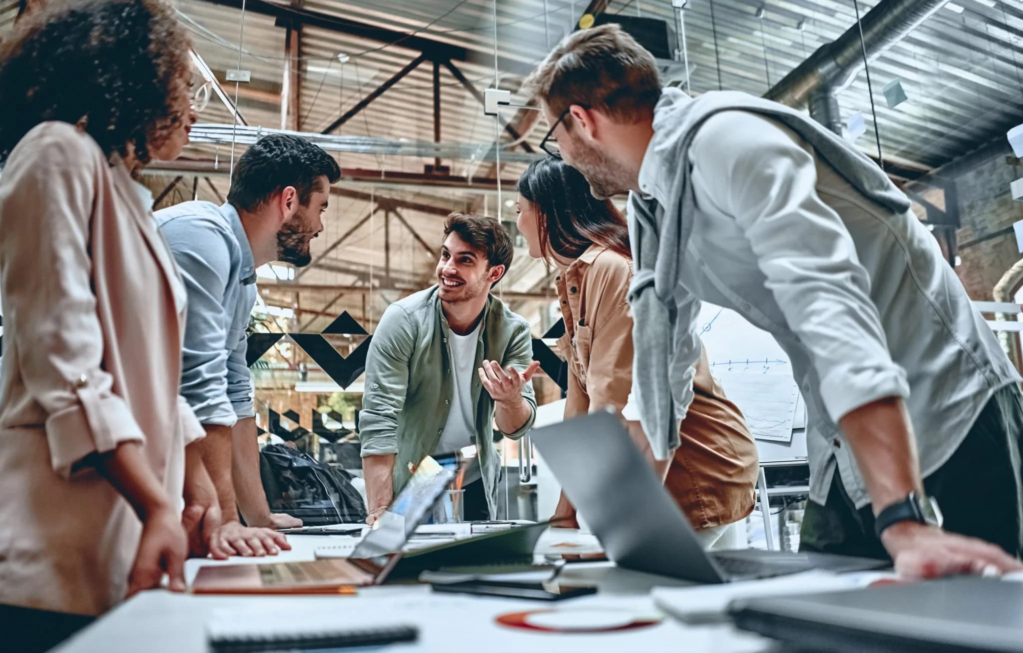 A group of colleagues gather around a table in a modern office, leaning in and actively discussing ideas, with laptops and documents spread out in front of them.