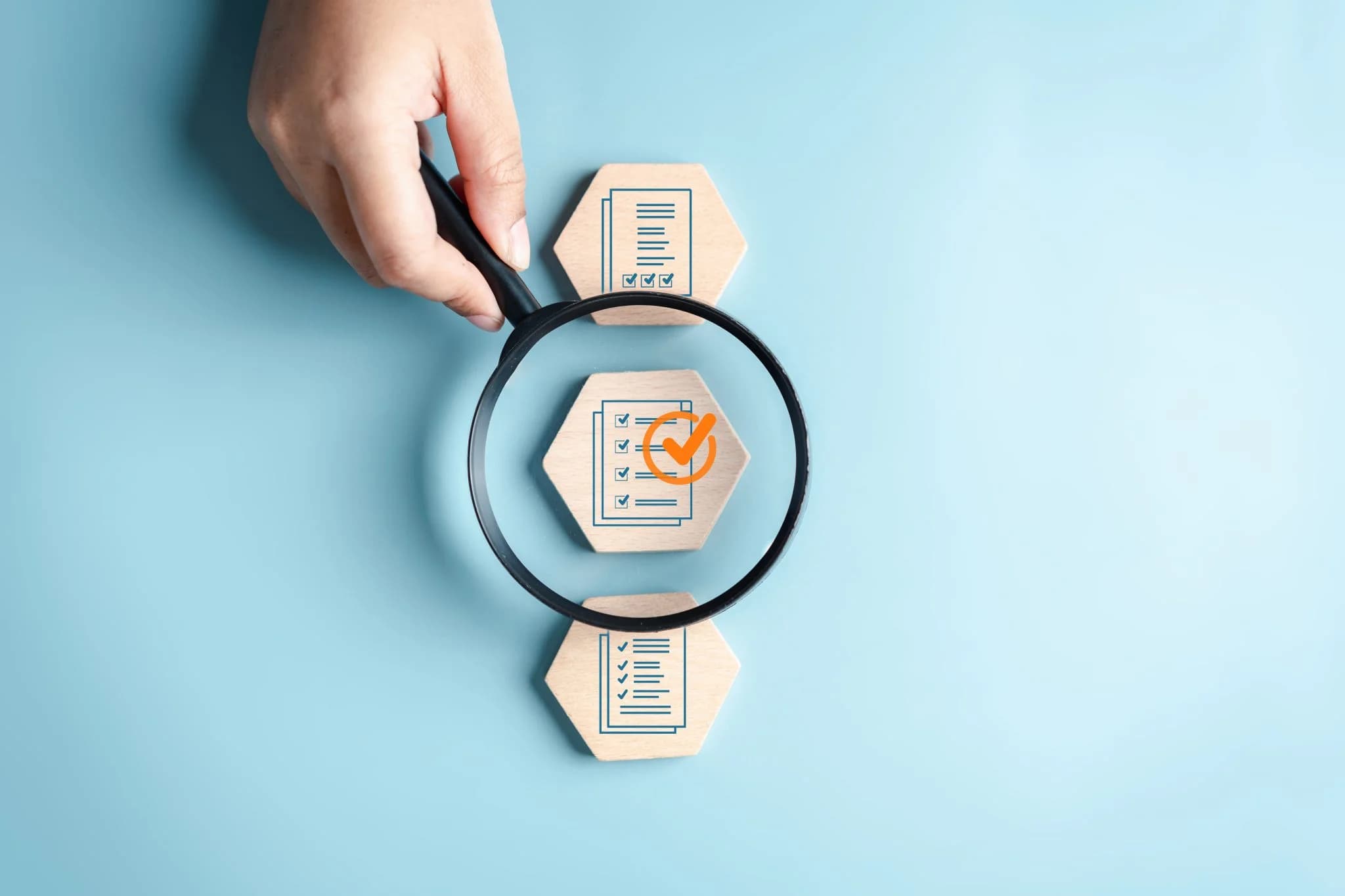 A hand holds a magnifying glass over wooden hexagonal tiles with checklist icons, highlighting one marked with a large orange checkmark