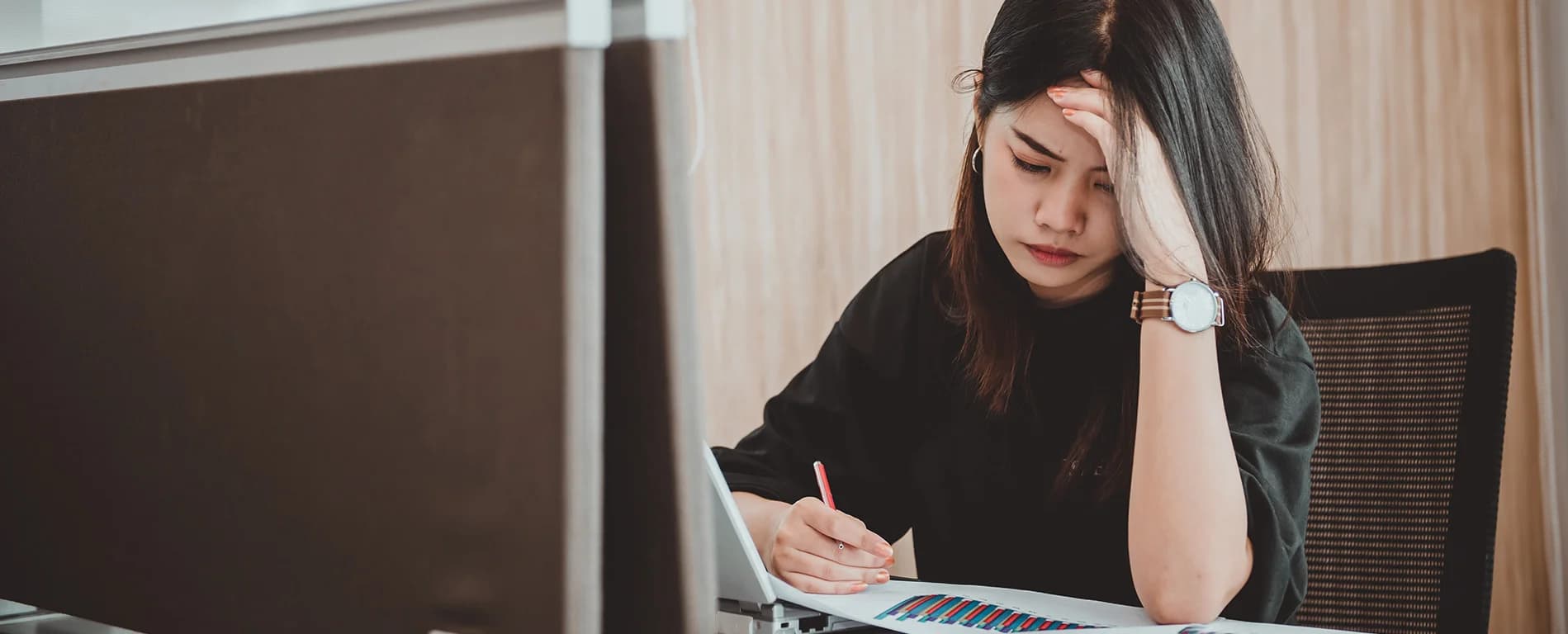 A woman sits at a desk looking stressed, holding her head while reviewing a printed chart next to a laptop.