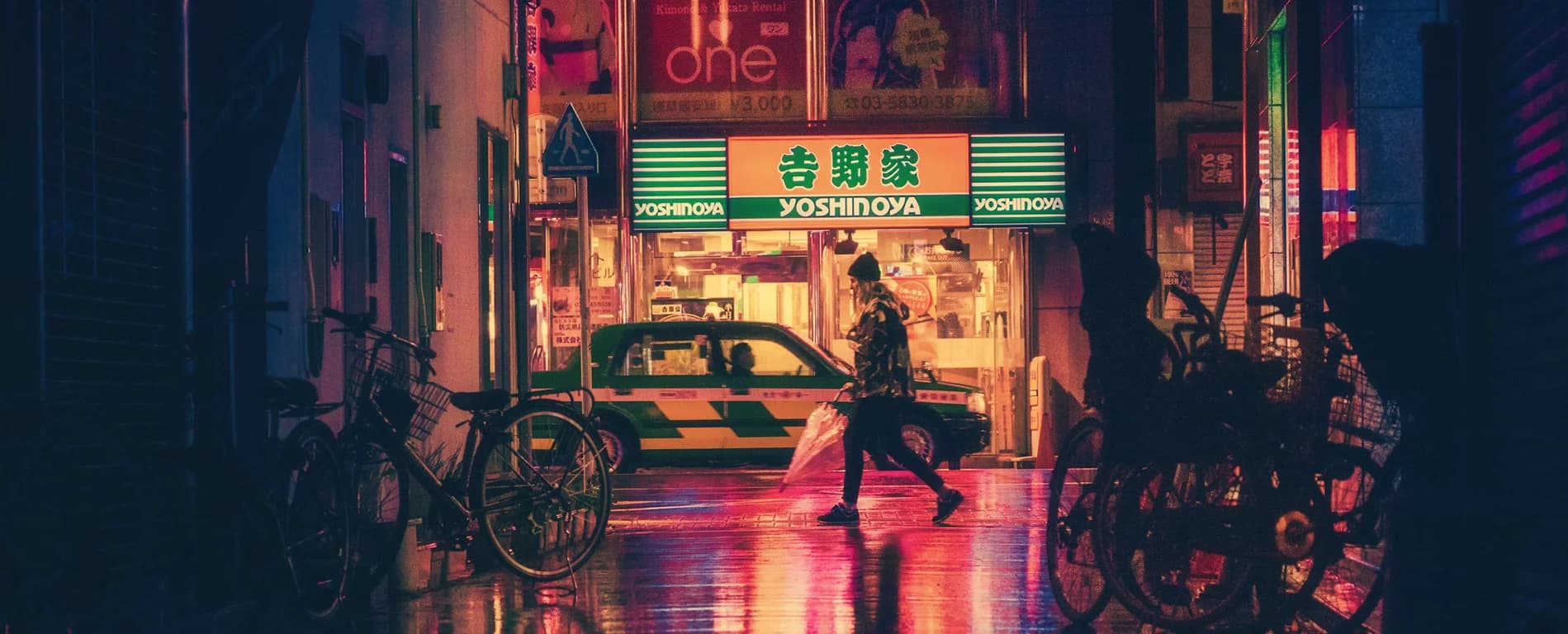A neon-lit urban street at night with a Yoshinoya restaurant storefront in the background. A person carrying an umbrella walks past a taxi, while bicycles line the sides of the narrow alley, reflecting colorful lights on the wet pavement.