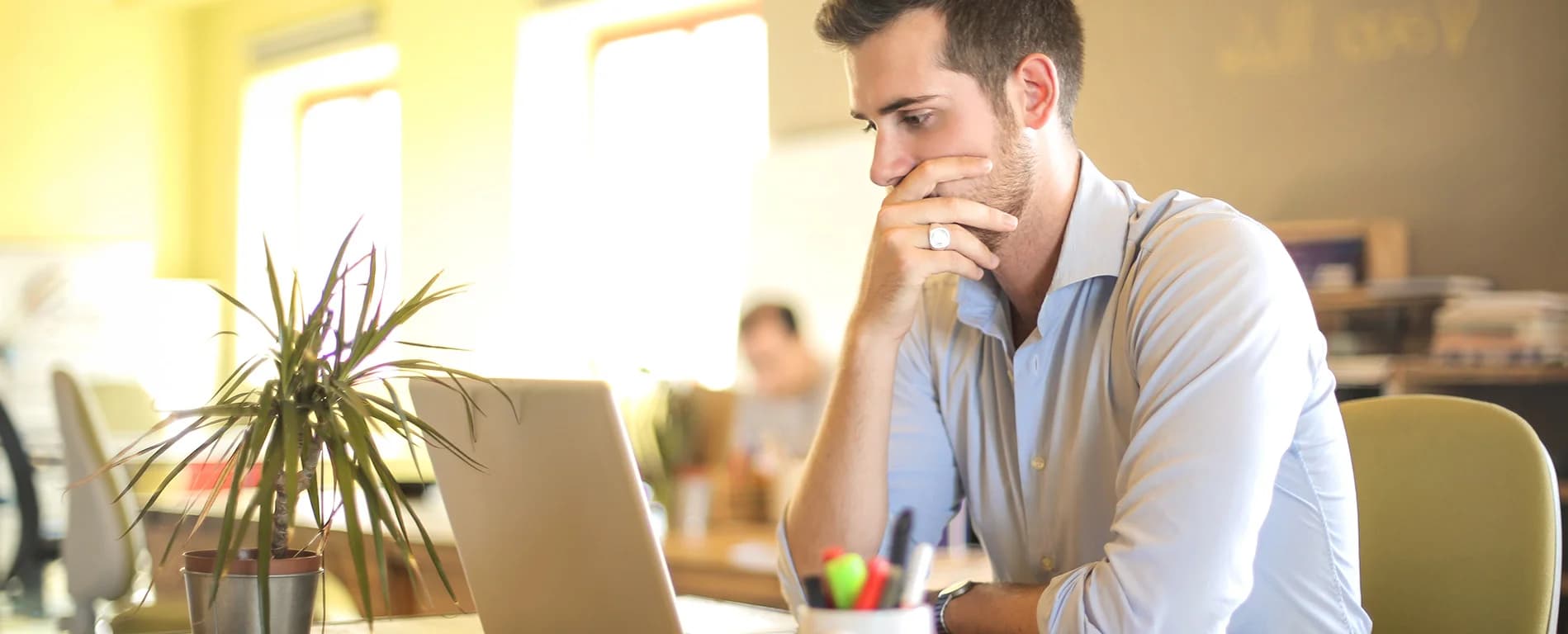 A man sits at a desk in a bright office, looking intently at a laptop screen with his hand covering part of his mouth in a thoughtful pose. A small potted plant and a cup holding colorful pens are on the desk in front of him. Sunlight streams through large windows behind him, and another person is blurred in the background, suggesting a shared workspace.