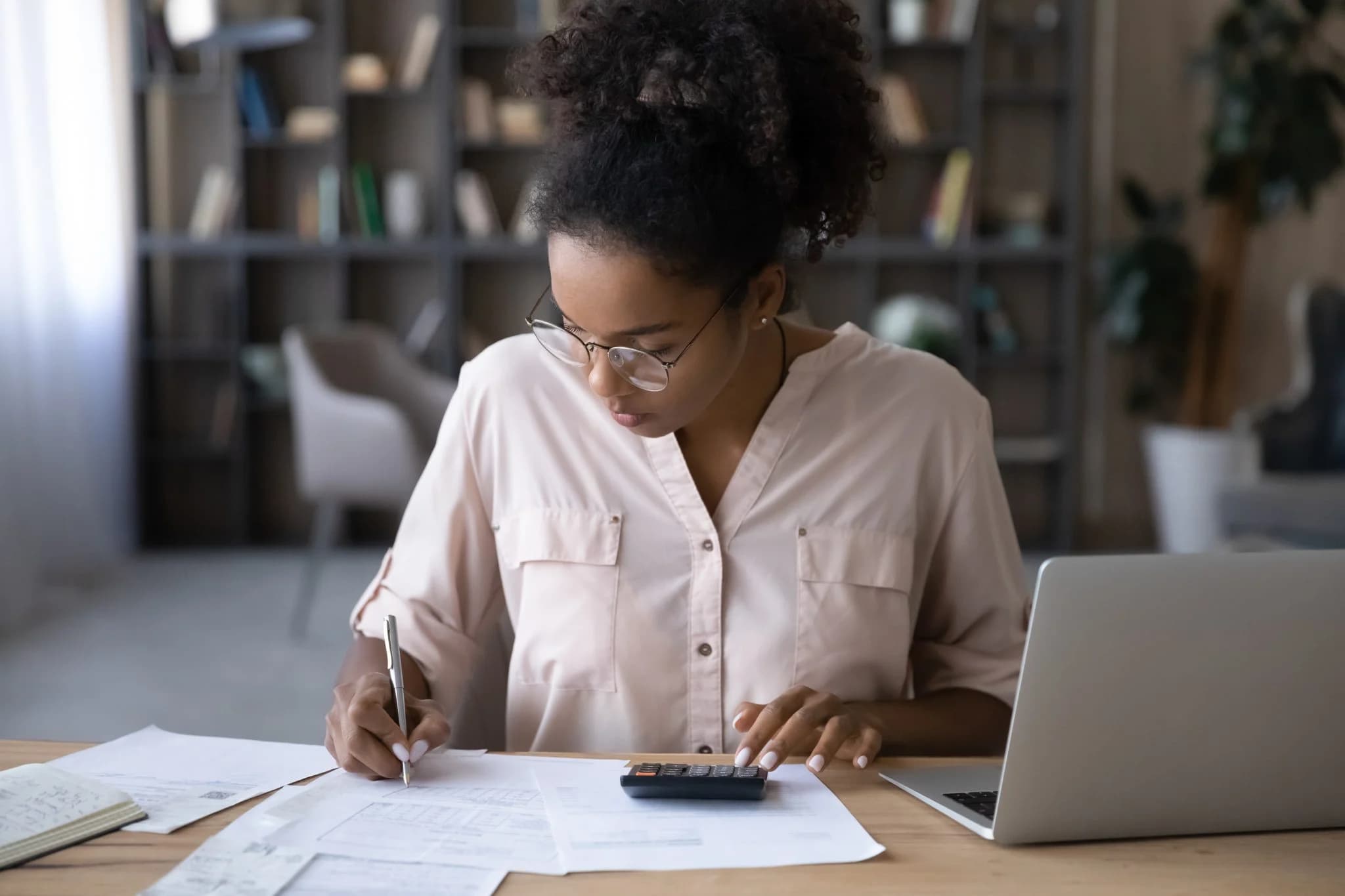 Woman calculating expenses at desk with documents, calculator, and laptop.