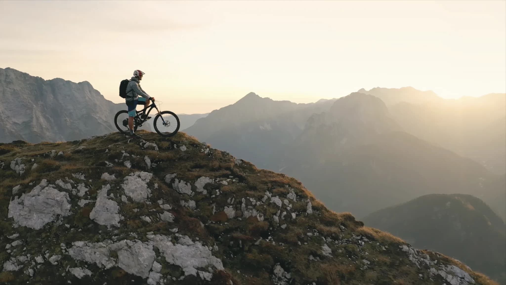 Mountain biker standing with a bike on a rocky peak at sunset, overlooking a mountain landscape.