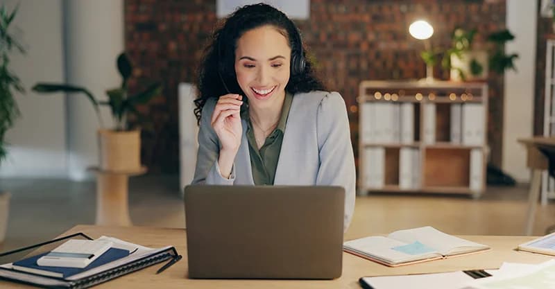 Smiling woman wearing a headset working on a laptop at a desk with notebooks in a home office.