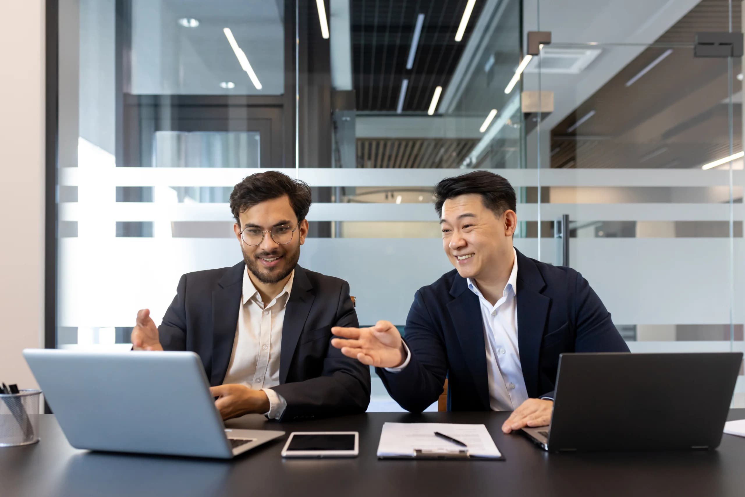 Two businessmen collaborating at a desk with laptops in a modern office.