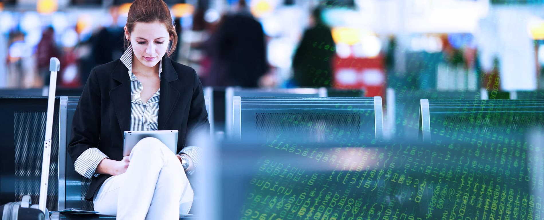Businesswoman sitting in an airport waiting area using a tablet with a suitcase beside her.