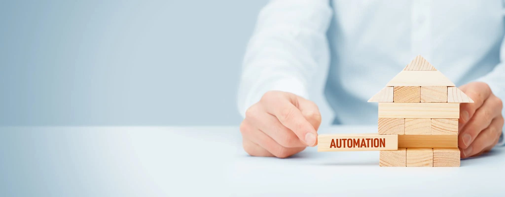 Hand placing a wooden block labeled "automation" into a small house made of wooden blocks