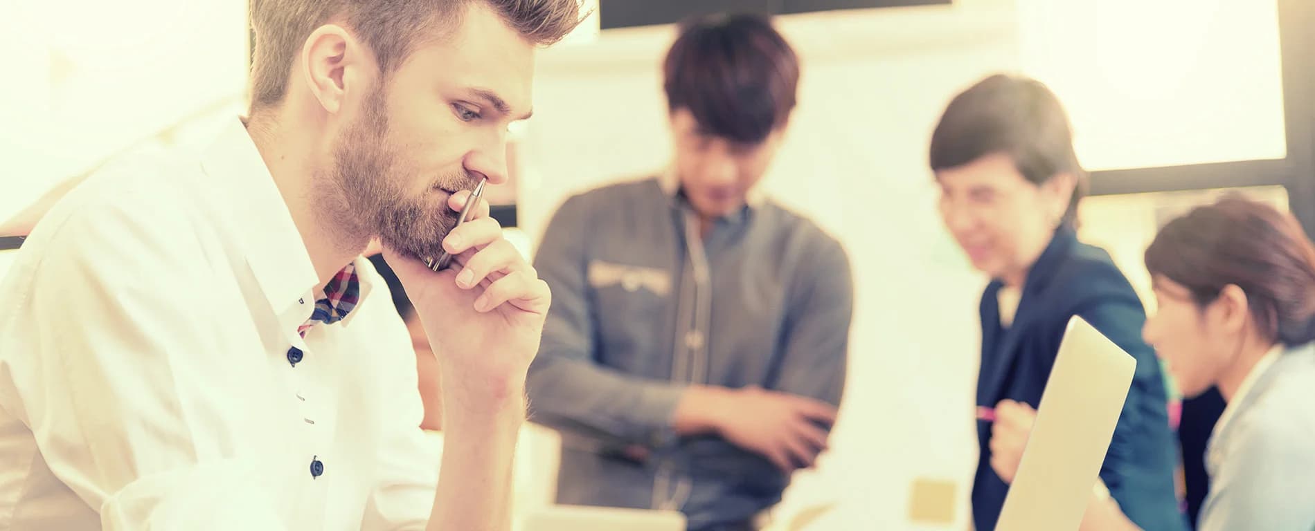 Man thinking while working on a laptop with colleagues in the background
