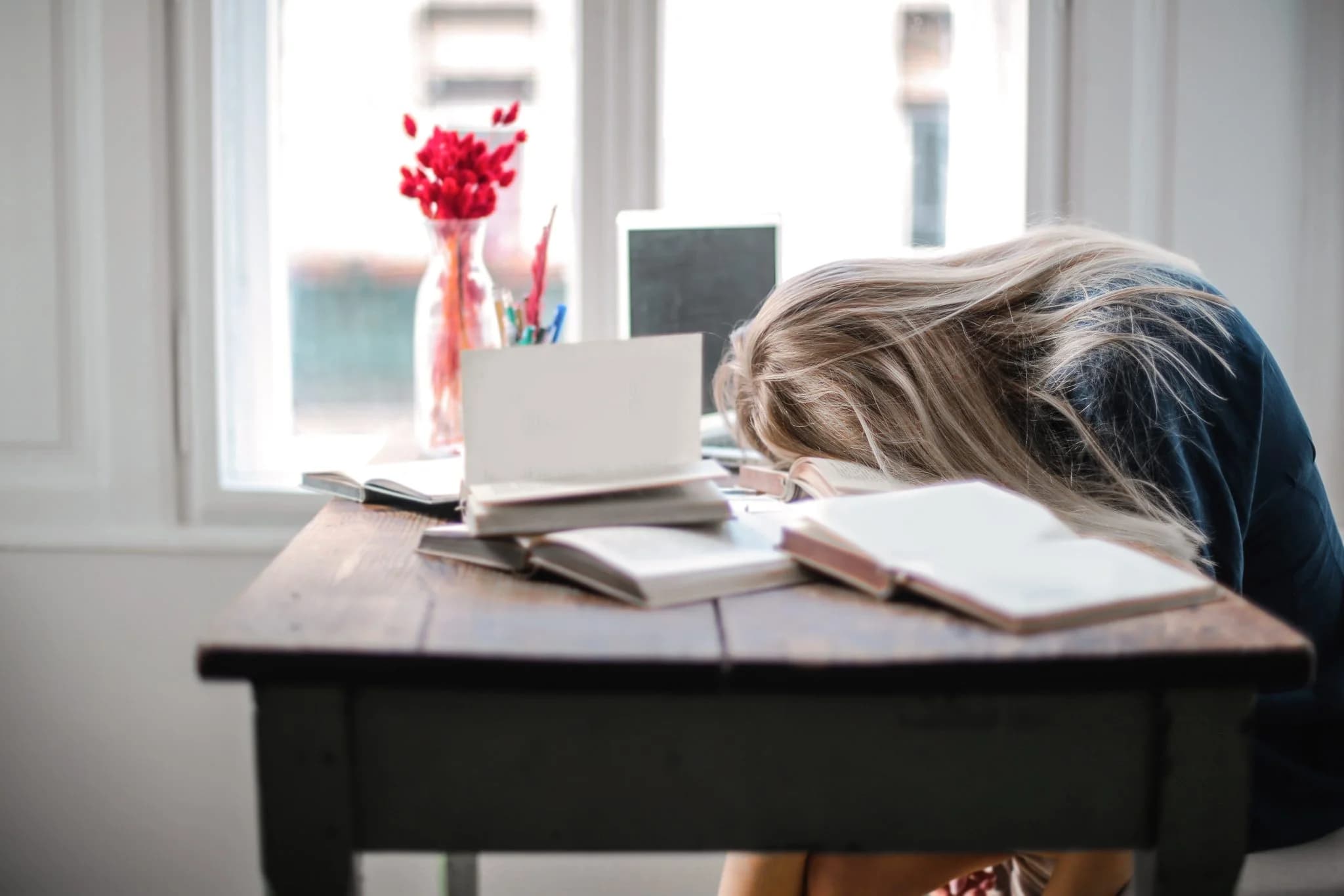 Person resting head on a desk with books, appearing tired or overwhelmed