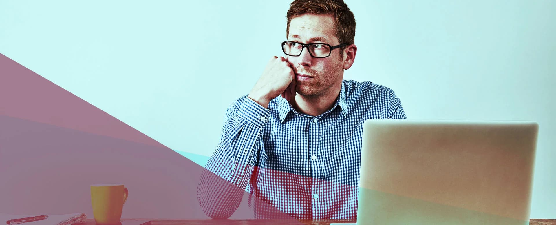 Man wearing glasses sitting at a desk, looking thoughtful while using a laptop.