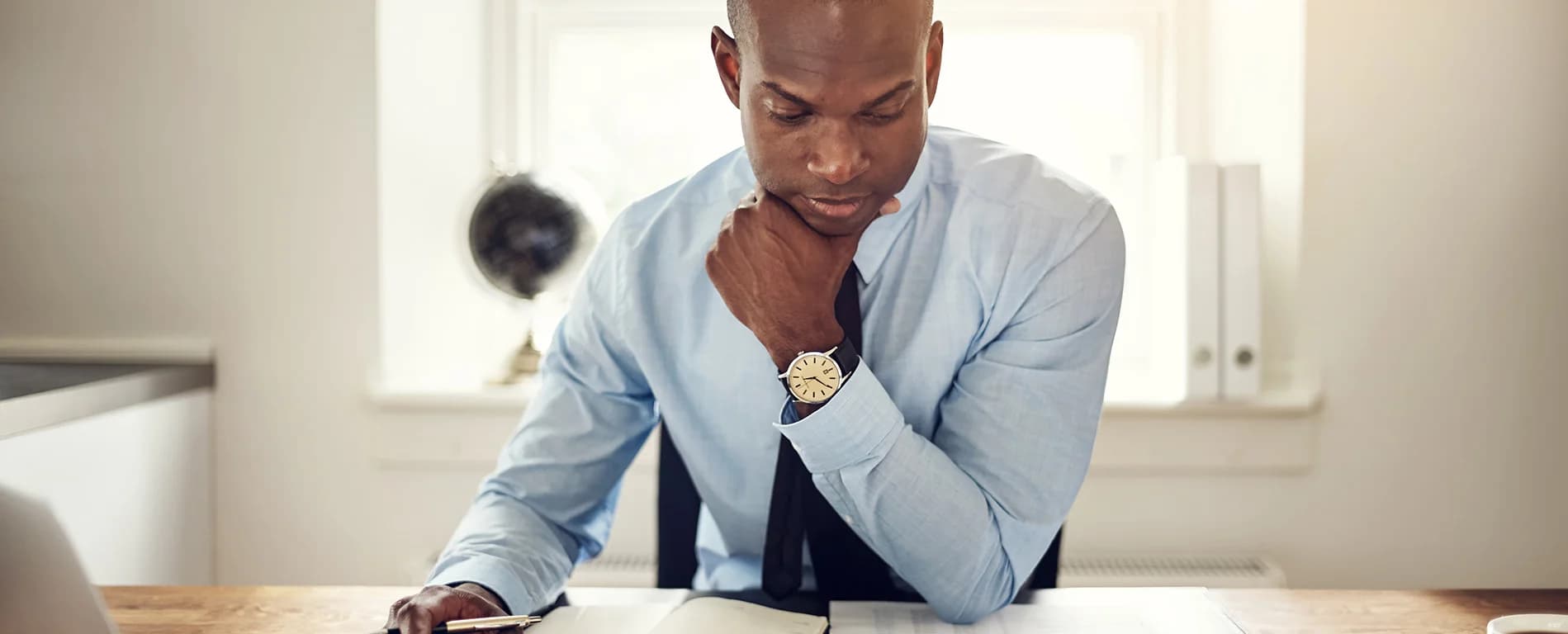 Man in a shirt and tie reading documents at a desk with a thoughtful expression.