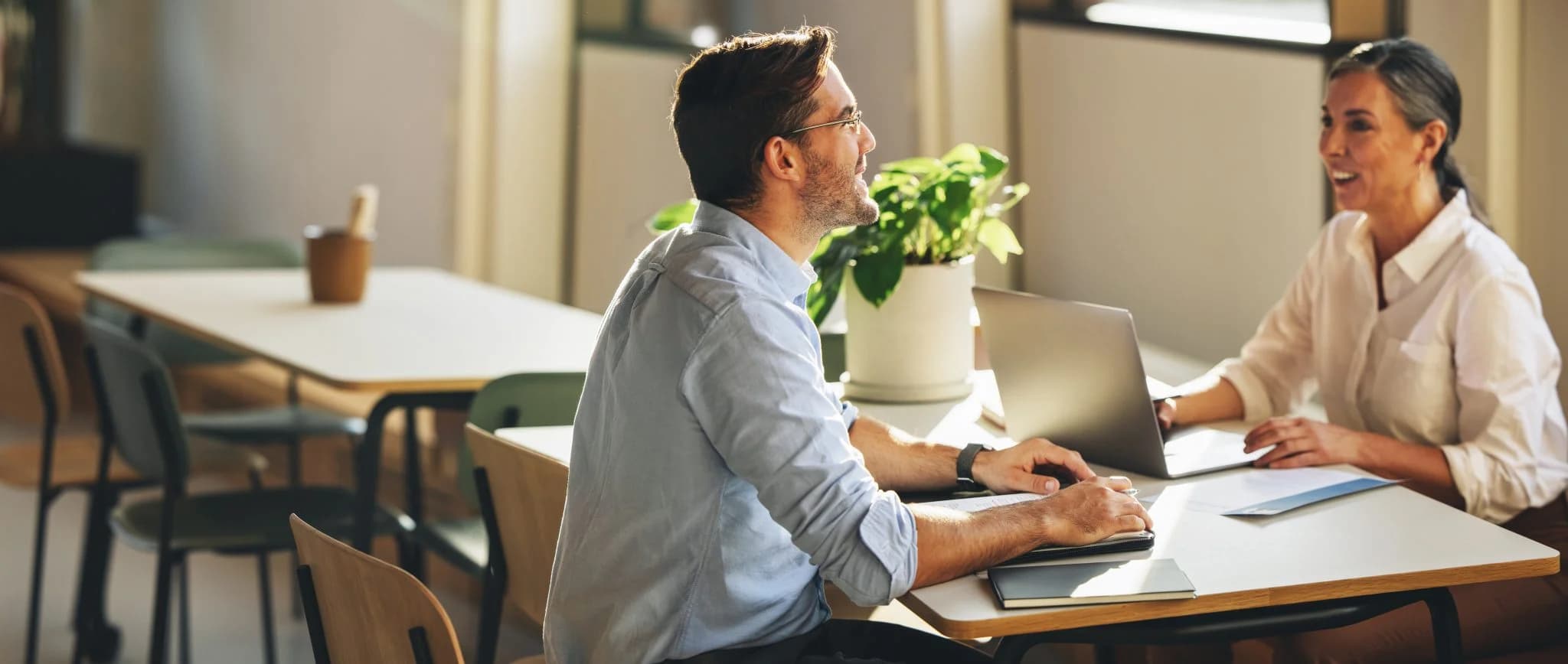 Two colleagues having a conversation while working on laptops at a table.