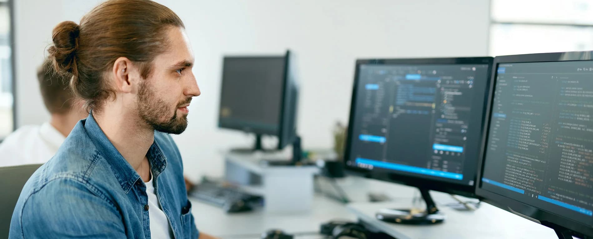 A man with his hair tied back in a bun works at a desk, looking at two computer monitors displaying lines of code in an office setting.