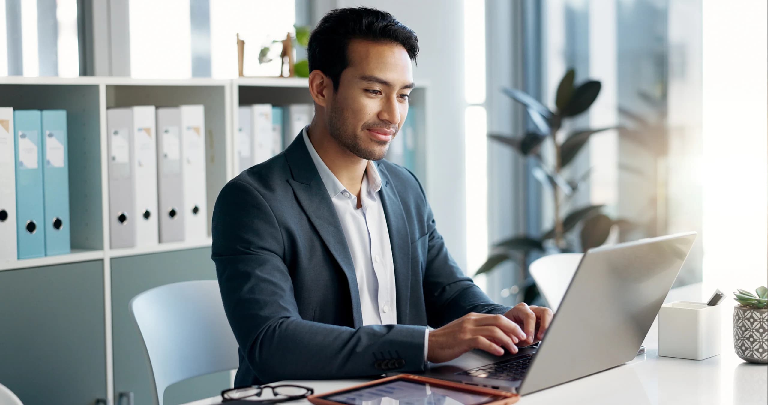 A professionally dressed man in a blazer sits at a desk in a bright office, typing on a laptop with folders and a plant visible in the background.