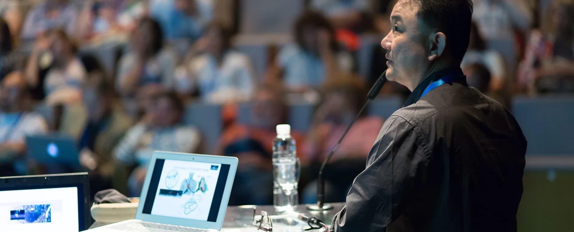 A man speaks into a microphone at a conference, standing beside a table with a laptop and water bottle, while an audience listens in the background.